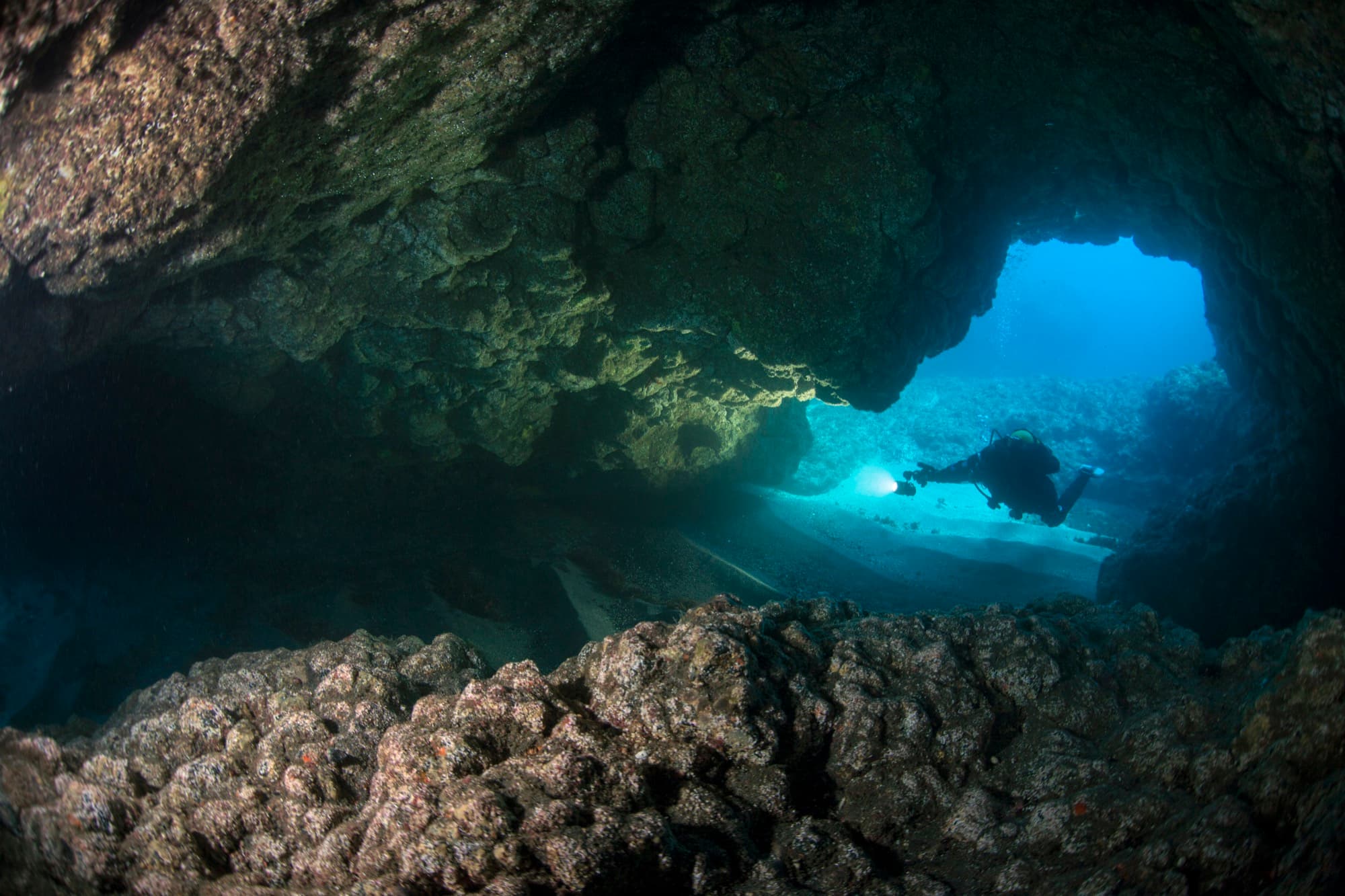 A diver with a headlamp beam exploring a sandy underwater cave passage